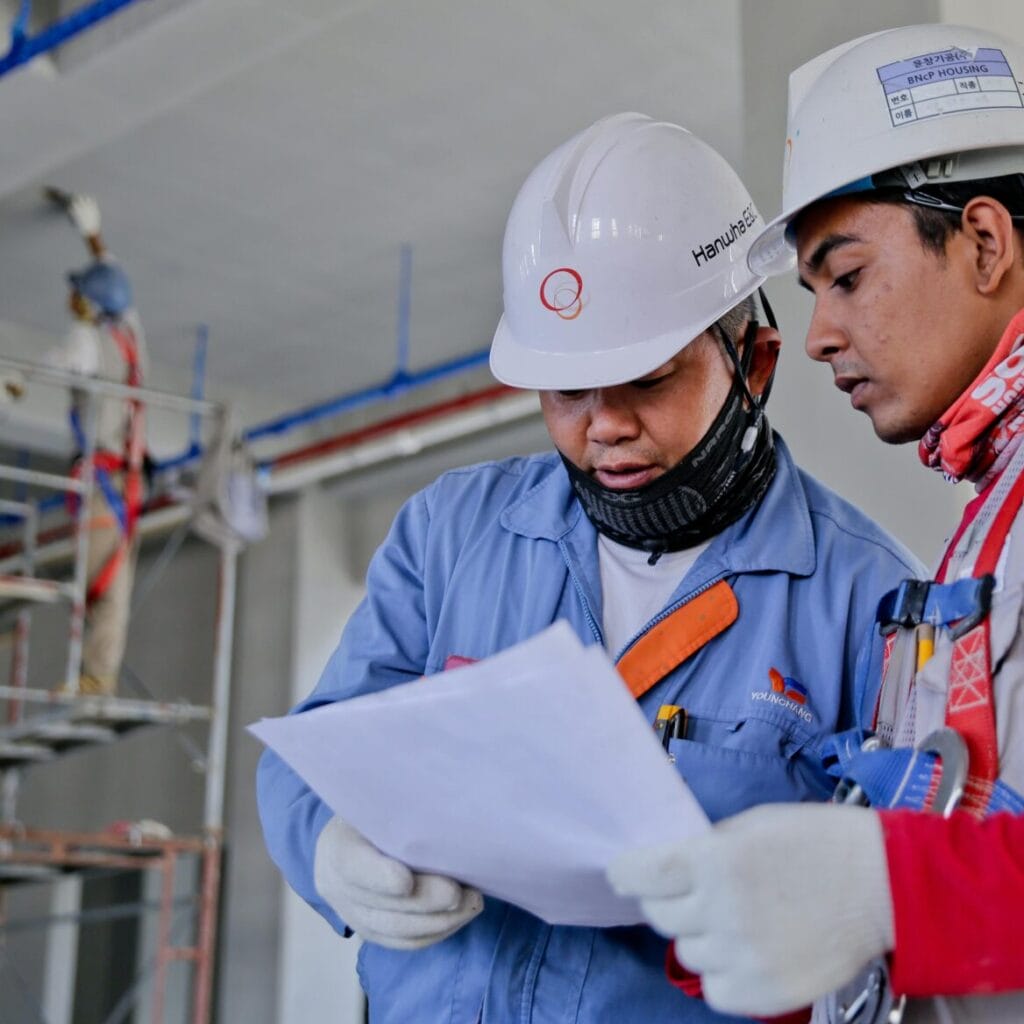 Two engineers in safety helmets reviewing construction plans at a worksite.